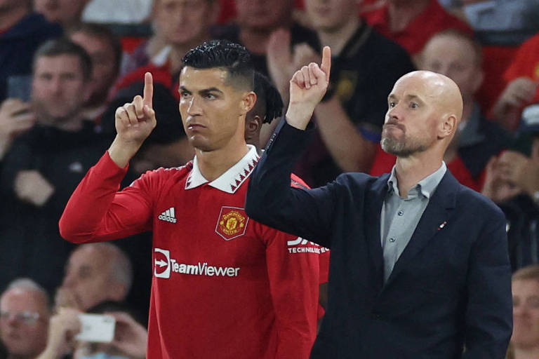 Cristiano Ronaldo e o treinador Erik ten Hag, do Manchester United, lado ao lado, apontam o dedo indicador direito para o alto em partida contra o Liverpool no estádio Old Trafford; atrás dele, torcedores acompanham o jogo