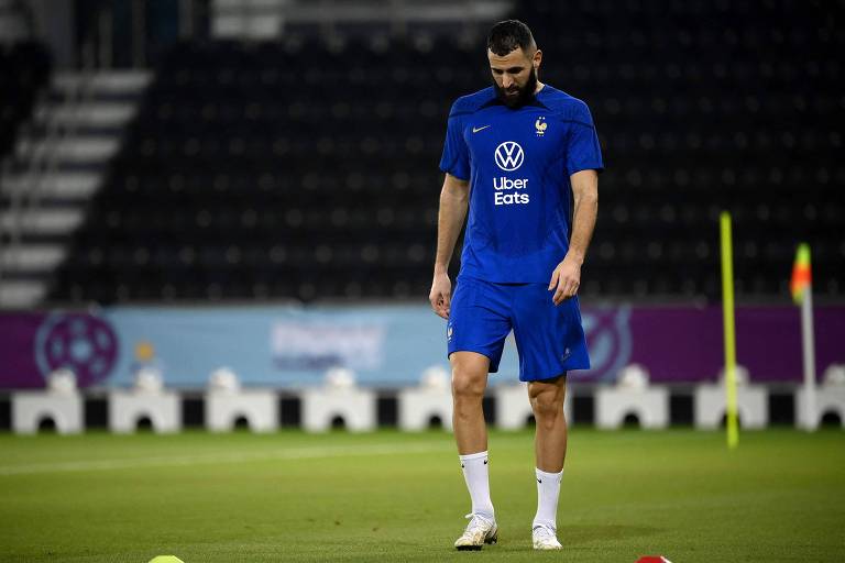 Com uniforme azul e meias brancas, o atacante Benzema, de cabeça baixa, anda em campo de treino da França em Doha