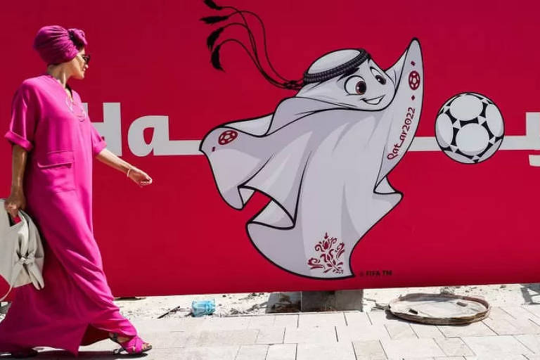 Imagem colorida mostra a mascote La'eeb, símbolo da Copa do Qatar; ele tem a forma de um lenço usado pelas pessoas do mundo árabe. Está pintado de branco em uma parede com fundo vermelho.