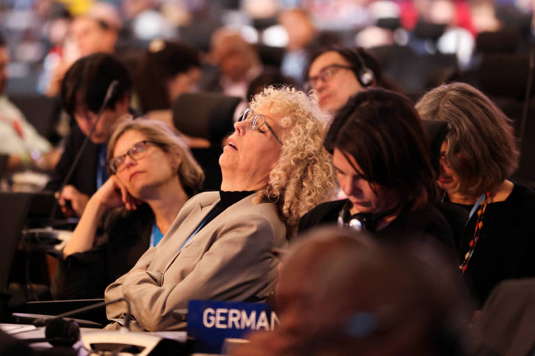 Mulher loira dorme sentada; na sua frente há uma placa dizendo Germany (Alemanha) e, ao seu lado, há mais pessoas que parecem também dormir