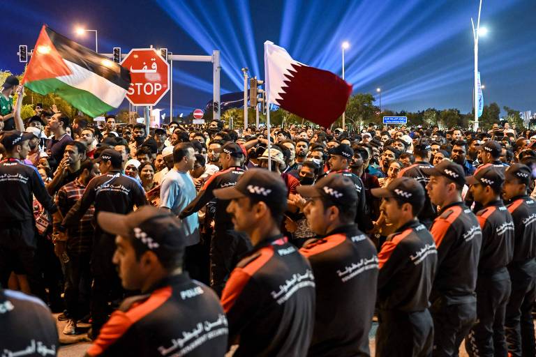 Lado a lado, policiais formam um cordão humano para evitar invasão de torcedores na Fan Fest no parque Al Bidda, em Doha, antes do jogo de abertura da Copa do Mundo do Qatar; diante deles há uma multidão de pessoas aglomeradas