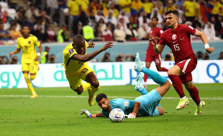 Imagem colorida mostra o goleiro qatariano, de uniforme azul, esticado no chão, com a mão direita tocando o equatoriano Valencia, que está correndo e se desequilibra.