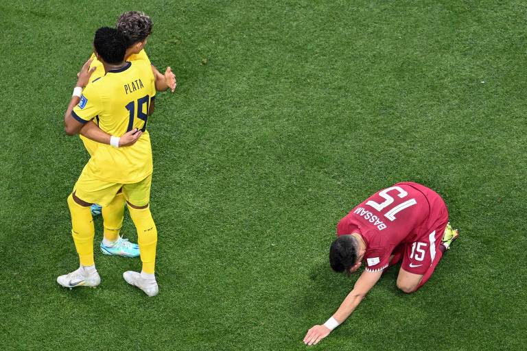 Imagem colorida mostra um jogador do Qatar, de uniforme vermelho, agachado, com o rosto encostado no gramado e a mão esquerda estendida; no lado esquerdo, dois jogadores do Equador, usando uniforme amarelo, estão em pé se abraçando.