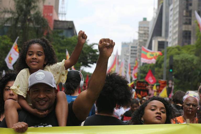 A imagem mostra uma manifestação em uma cidade, onde um homem carrega uma menina em seus ombros. A menina, com cabelo cacheado e vestido claro, levanta o punho em sinal de protesto. O homem, com uma camiseta escura e boné, também levanta o punho. Ao fundo, há várias pessoas e bandeiras coloridas, além de prédios altos e árvores.