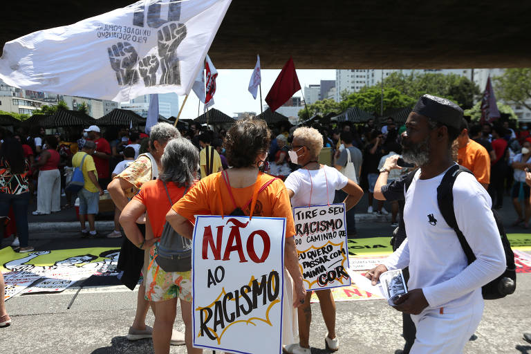 A imagem mostra um grupo de pessoas participando de um protesto. Algumas pessoas estão segurando cartazes， incluindo um que diz 039;NÃO AO RACISMO039;. Há bandeiras brancas e vermelhas ao fundo， e a multidão parece estar reunida em um espaço público， com árvores e edifícios visíveis ao fundo.