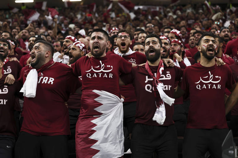 Vários homens abraçados cantando e vestindo camisetas na cor vinho escritas Qatar