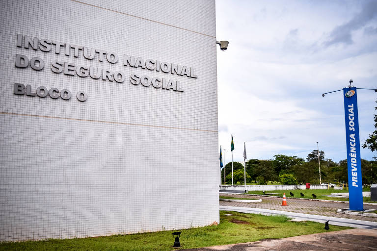 Foto mostra a fachada do INSS em Brasília. Em primeiro plano vemos um muro onde o nome do institudo está grafado em letras prateadas. 