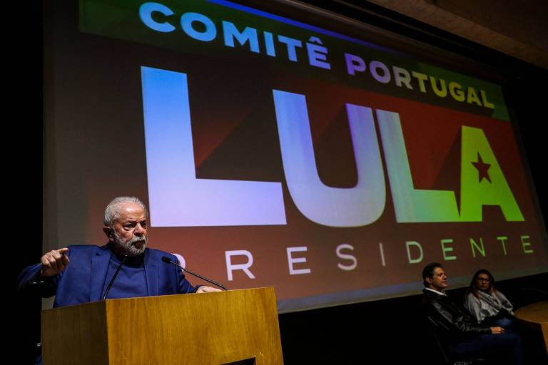 Lula fala em púlpito de madeira com telão ao fundo onde se lê em letras brancas sobre fundo vermelho "Comitê Portugal Lula Presidente". No canto direito da foto, bem pequenos, estão Fernando Haddad e Janja, sentados