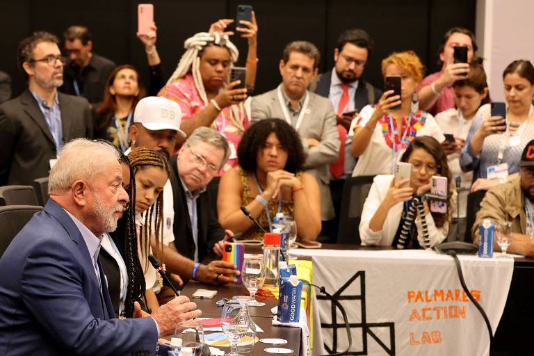 Lula fala sentado a uma mesa com muitas pessoas; outras mais, de pé, se esticam para enxergar e batem fotos com o celular