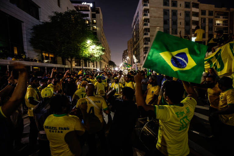 torcedores de verde e amarelo se aglomeram em rua do Qatar, à noite. a bandeira do Brasil está na mão de um deles, logo em primeiro plano