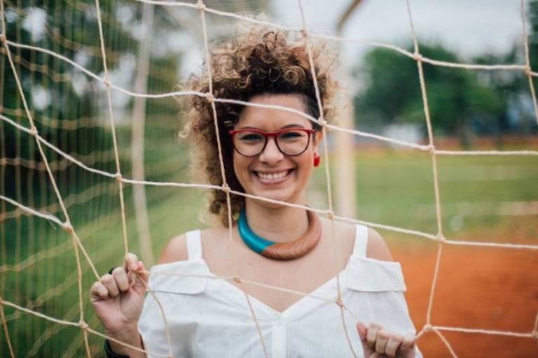 aira bonfim, mulher branca de cabelos cacheados, com luzes, é fotografada atrás de uma rede de futebol. ela está sorrindo