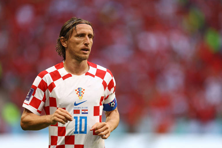 O jogador croata Modric está com a camisa da seleção croata, que é xadrez e branca e vermelha, durante jogo da Copa contra o Marrocos; ao fundo, os torcedores do time estão desfocados