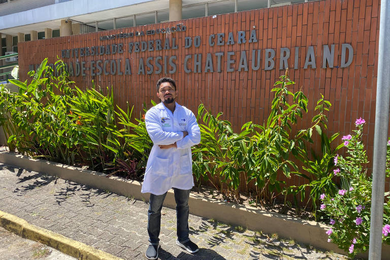 Um médico, um homem negro de jaleco, cruza os braços em frente a hospital onde lê-se Maternidade Escola Assis Chateaubriand, Fortaleza, Ceará