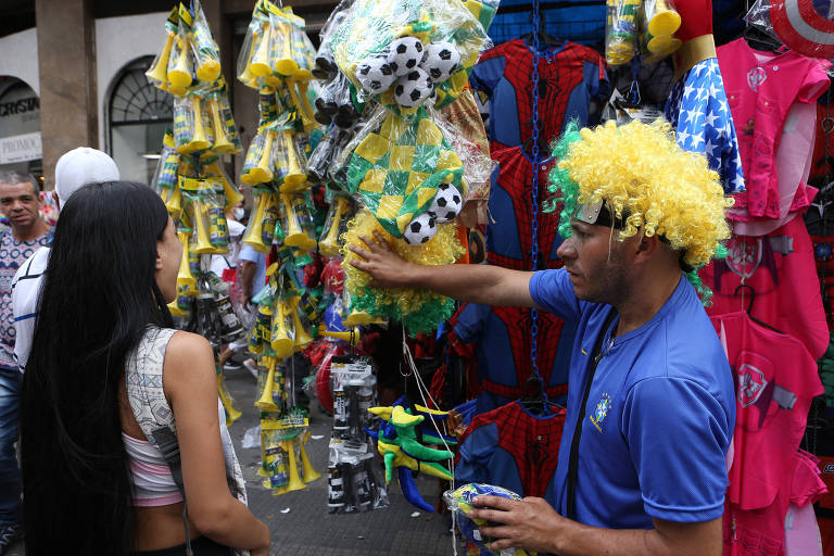 Ambulante mostra mercadorias para consumidora. Sacolas penduradas estão cheias de produtos com as cores do Brasil. Ao lado, há fantasias.