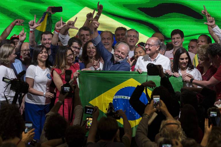 Lula, homem branco, está no centro, cercado de pessoas e atrás de uma bandeira do Brasil estendida