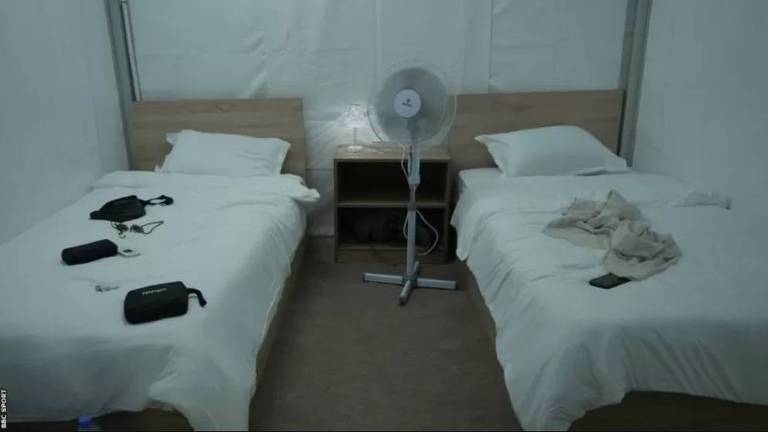 Parte de dentro de uma tenda com duas camas, uma mesinha e um ventilador no meio