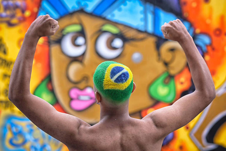 Jovem de costas, com os braços levantados; seu cabelo está pintado com a bandeira do Brasil