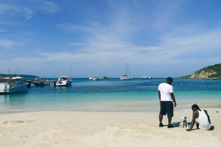 Em primeiro plano, um homem em pé e outro sentado na areia, ambos de camiseta branca; ao fundo o mar verde água com alguns barcos