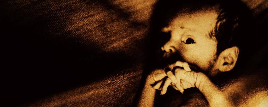 Retrato de bebê ianomâmi em rede; ele olha para a câmera e tem os dedos das mãos cruzados