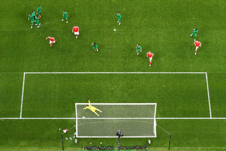 Jogadores de Rússia (de camisas vermelhas) e da Arábia Saudita (de uniforme verde) observam a bola entrar no gol depois de cobrança de falta de Golovin em Moscou, na Copa de 2018; o goleiro, vestido de amarelo, pula mas não alcança a bola
