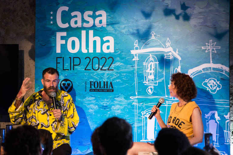 Homem de amarelo fala a uma platéia, ao lado de uma mediadora ruiva; no painél atrás dele lê-se Casa Folha - Flip 2022