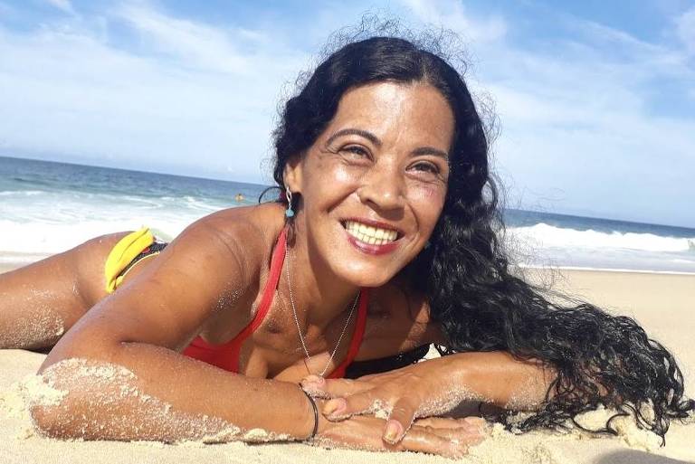 mulher negra, cabelos pretos cacheados longos, posa para foto deitada na areia da praia, é possível ver as ondas do mar atrás dela. Está sorrindo