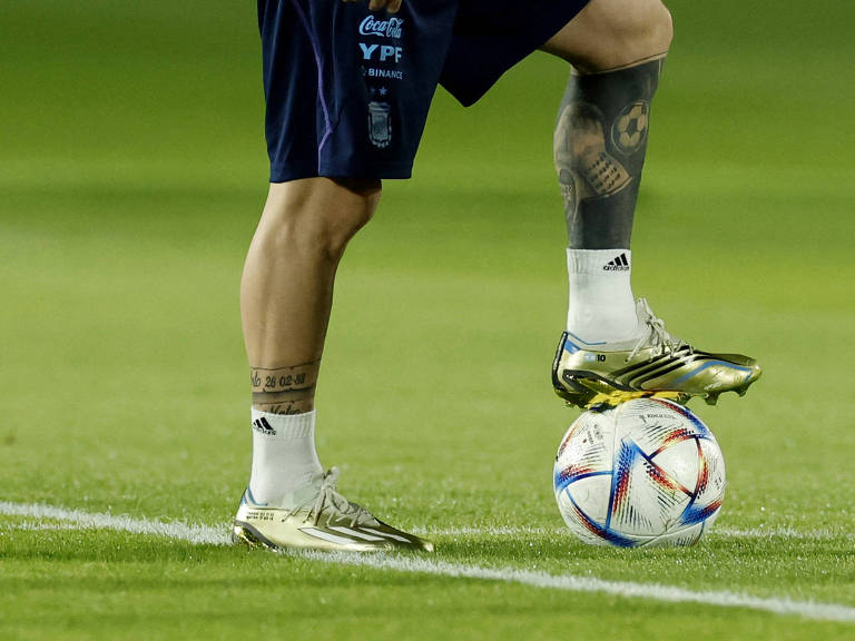 Imagem mostra as pernas de argentino Messi em treino no Qatar. Ele está de pé, e um de seus pés, o esquerdo, pisa sobre uma bola. O jogador usa chuteiras prateadas e calção azul escuro