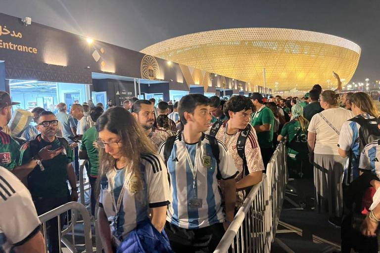 Imagem colorida feita à noite mostra vários torcedores com camisas da Argentina, brancas com listras azul claro, e do México, na cor verde. Ao fundo, um estádio em forma de concha está iluminado.
