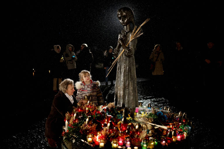 Uma mulher e criança são vistas colocando alimentos e enfeites ao lado da estátua de uma criança, que representa uma vítima da grande fome na Ucrânia/ pequenas velas iluminam a estátua, que está na penumbra
