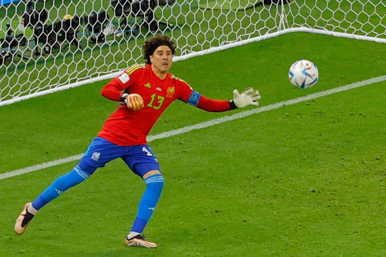 Imagem colorida mostra o goleiro Ochoa, de corpo inteiro, saltando para tentar pegar a bola na direita. Ele veste camisa vermelha e calção azul. Tem cabelo volumoso, preso com uma faixa.