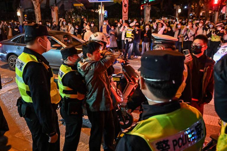 Em uma rua cercada de pessoas com máscaras, um homem entra em confontro com policiais que usam coletes amarelos

