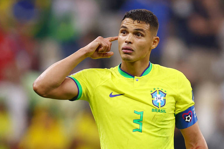 De camisa amarela da seleção com o número 3 em verde no peito, Thiago Silva, com a tarja azul de capitão do Brasil no braço esquerdo e olhando sem sorrir para a frente, leva o dedo indicador da mão direita à orelha direita no jogo de estreia na Copa do Mundo do Qatar, em Lusail, contra a Sérvia