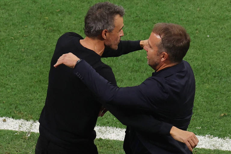 Imagem colorida mostra os técnicos de Espanha e Alemanha, da cintura, se abraçando. Os dois vestem roupa preta e estão de lado.