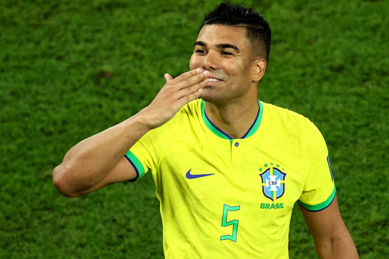 Imagem colorida mostra Casemiro, da cintura para cima, vestindo a camisa amarela da seleção brasileira, com o número cinco na frente. Ele está sorrindo, com a mão direita na boca, como se estivesse mandando um beijo.