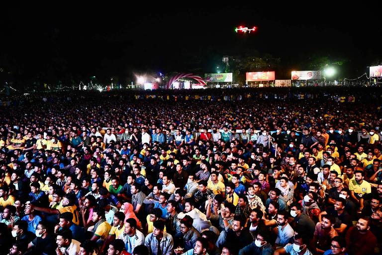 Imagem colorida, registrada em um local aberto e à noite, mostra centenas de pessoas sentadas