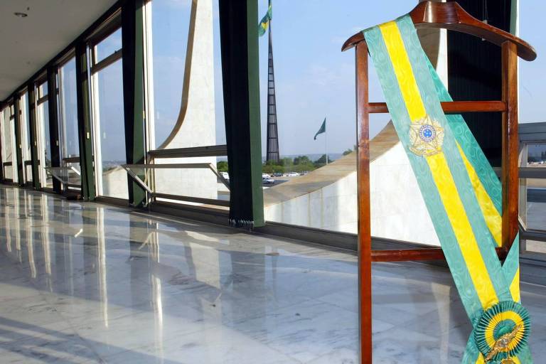 Foto mostra faixa presidencial verde e amarela em um cabide de madeira, instalado dentro do Palácio do Planalto. Ao fundo, as pilastras do Palácio e a bandeira brasileira hasteada