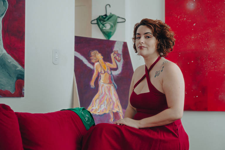 Um mulher ruiva de vestido vermelho aparece sentada com os braços cruzados no colo em um ambiente com um quadro vermelho ao fundo e sofá também vermelho