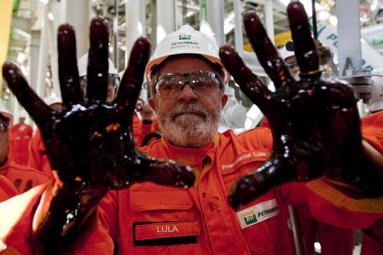 Lula de boné, óculos de proteção e macacão laranja da Petrobras mostra as duas mãos espalmadas, cobertas de óleo preto