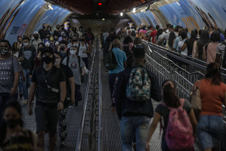 A imagem mostra uma estação de metrô movimentada, com muitas pessoas caminhando em direções opostas. A maioria dos indivíduos está usando máscaras. O ambiente é iluminado, com paredes decoradas e um teto curvo. À direita, há uma grade que separa os passageiros. A cena transmite uma sensação de agitação e movimento.