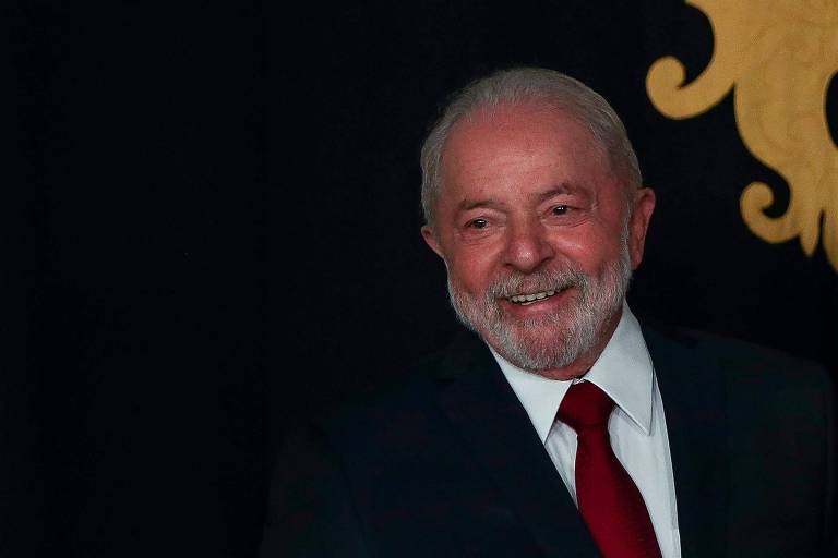 Lula, homem branco, visto do peito para cima, ocupando a metade direita da imagem. Está de camisa branca, paletó escuro e gravata vermelha. Fundo escuro com desenho em tom amarelo no canto superior direito