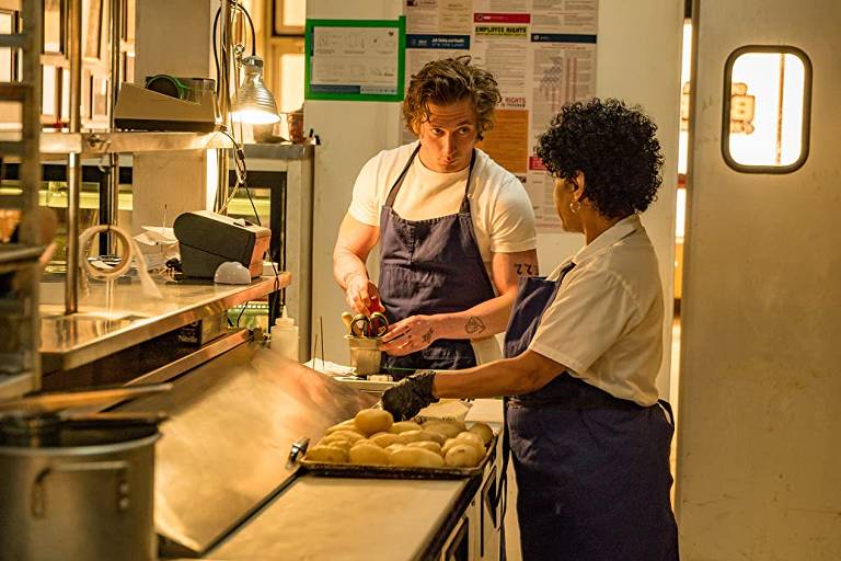 Um homem branco e uma mulher latina, ambos vestindo camisetas brancas e aventais azul-escuro, conversam em uma cozinha de restaurante. Ela tem uma bandeja cheia de batatas na sua frente e está de luvas de látex pretas.
