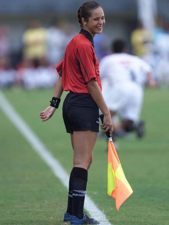 Com camisa vermelha e calções e meias pretos, Ana Paula Oliveira atua como bandeirinha em jogo beneficente na Vila Belmiro, em Santos, em 2003; na mão direita ela segura uma bandeira quadriculada nas cores amarelo e laranja