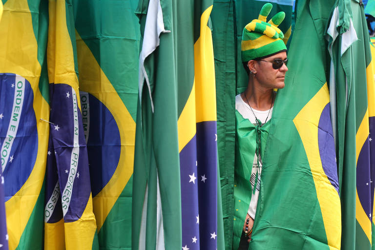 Imagem mostra varias bandeiras no Brasil hasteadas e um homem, com o corpo parcialmente coberto usando um oculos e um chameu com as cores do Brasil, aparece em meio as bandeiras
