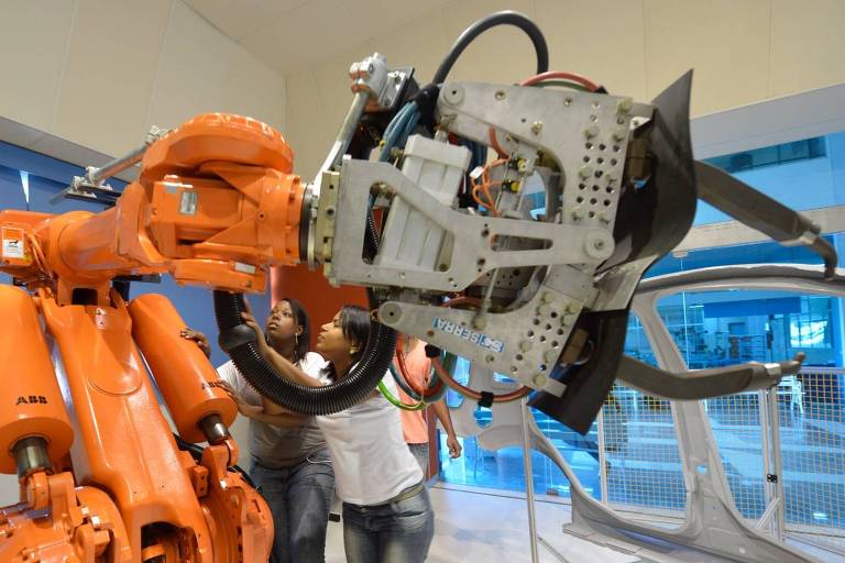 Imagem do braço de um robô em uma linha de montagem, sendo operado por uma funcionária