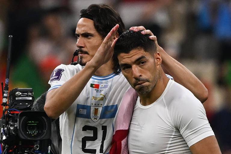 Cavani, com a camisa com o número 21, passa as mãos na cabeça de Suárez, colega de seleção uruguaia, depois da derrota para Portugal na Copa do Qatar; ao lado de Cavani há uma câmera de TV 