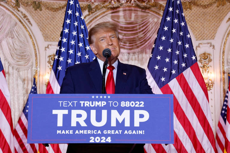 Donald Trump em púlpito; ele é um homem branco, loiro e veste terno preto com camisa branca e gravata vermelha; atrás dele, estão bandeiras dos EUA