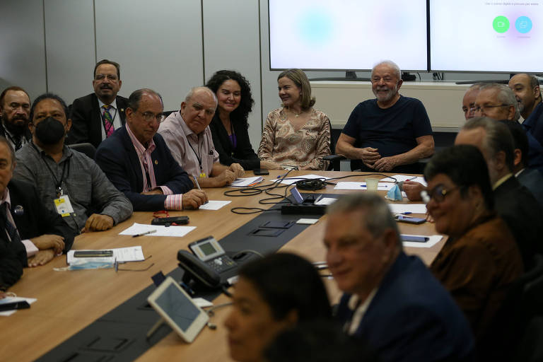 Presidente eleito Luiz Inácio Lula da Silva participa de reunião com representantes de centrais sindicais. Todos estão sentados a uma grande mesa, com Lula em uma das extremidades. Ele veste uma camiseta preta e aparece sorrindo