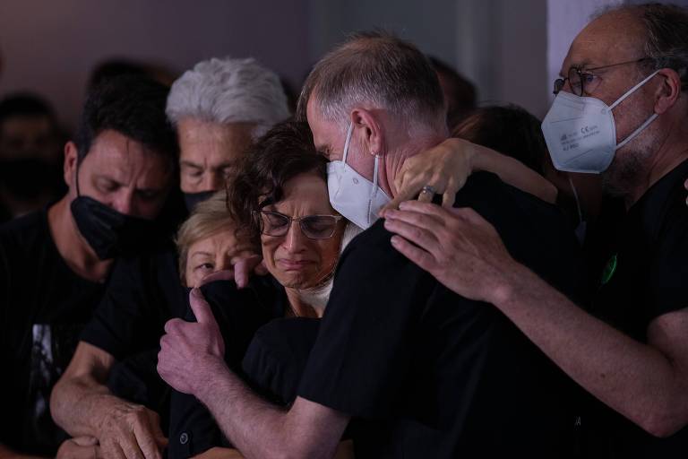 mulher no centro da imagem recebe abraço coletivo, de amigos e familiares, durante velório do marido; ela está de óculos e chora enquanto é reconfortada. Todos vestem preto e dois homens usam máscaras de proteção brancas. 