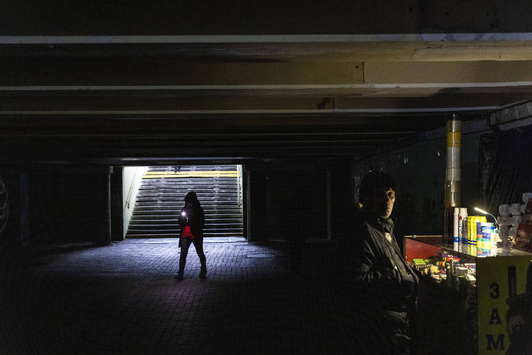 Mulher com lanterna passa por loja também iluminada por lanternas em passagem subterrânea em um bairro em Kiev