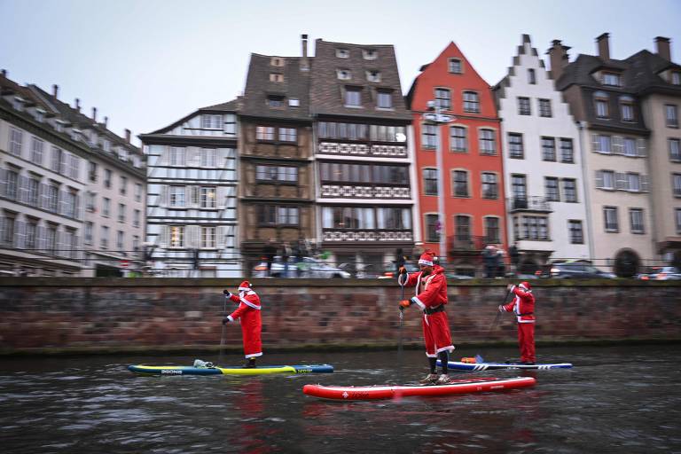 No primeiro plano, 3 pessoas vestidas de Papai Noel em Stand Up Paddles remam em um rio. No segundo plano, prédios típicos do leste da França, na beira do rio.